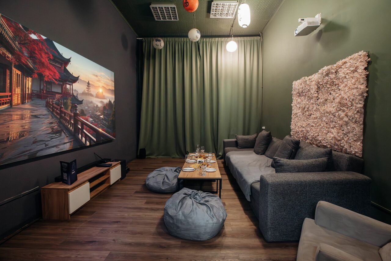 Арендовать Зал Китай. Лофт-зал в китайском стиле . Комната с темно-зелеными стенами и шторами, серая мягкая мебель, пара пуфов, деревянный пол, столик с посудой, декоративная стена из натурального материала, проектор, большой экран с изображением азиатского пейзажа, дисперсное освещение.