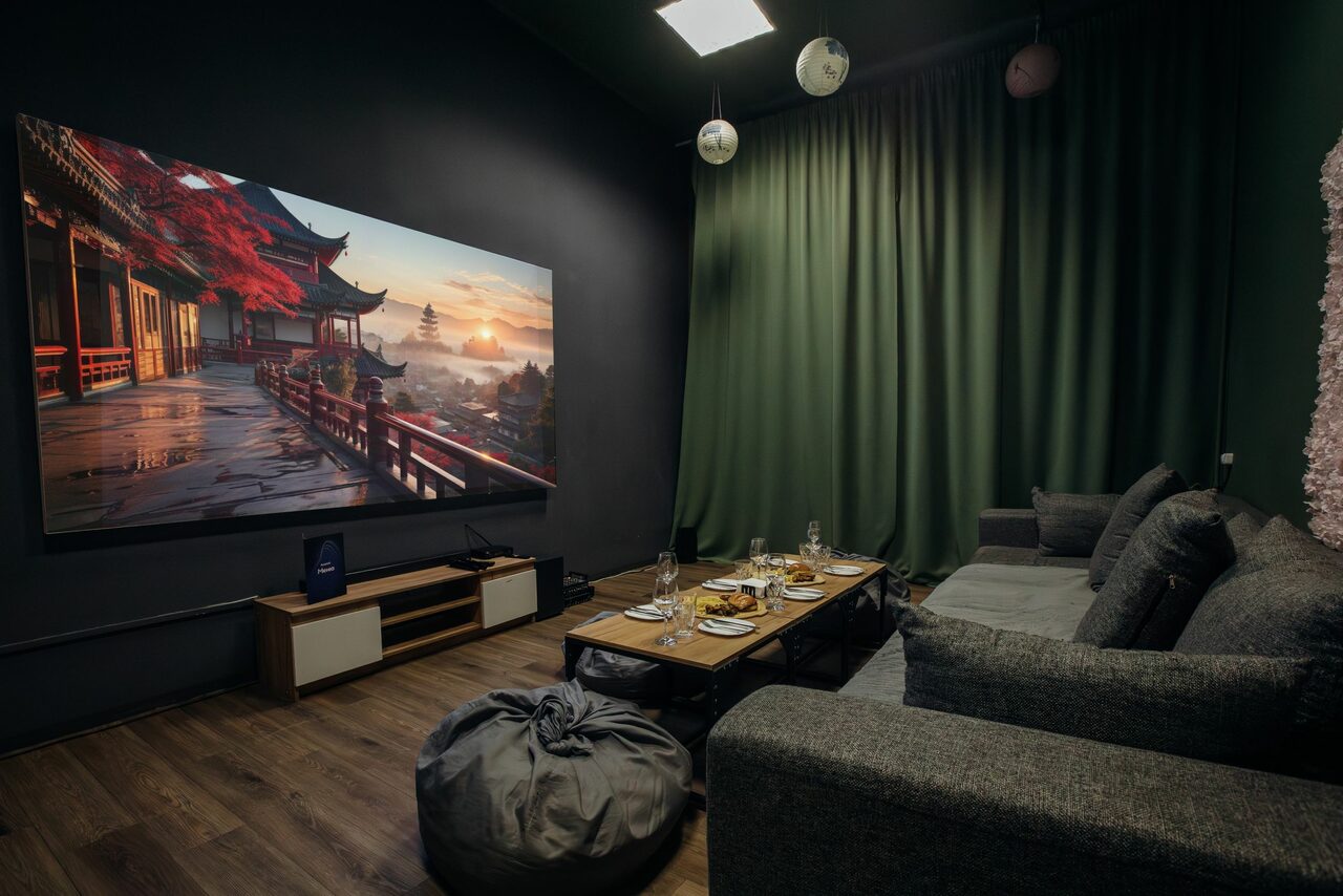 Арендовать Зал Китай. Лофт-зал в китайском стиле . Комната с деревянным полом, серыми стенами и зелеными шторами. Серый угловой диван, пуф, кофейный столик с сервировкой. Большой экран на стене, деревянная полка под ним. Спокойное освещение, потолочные светильники.