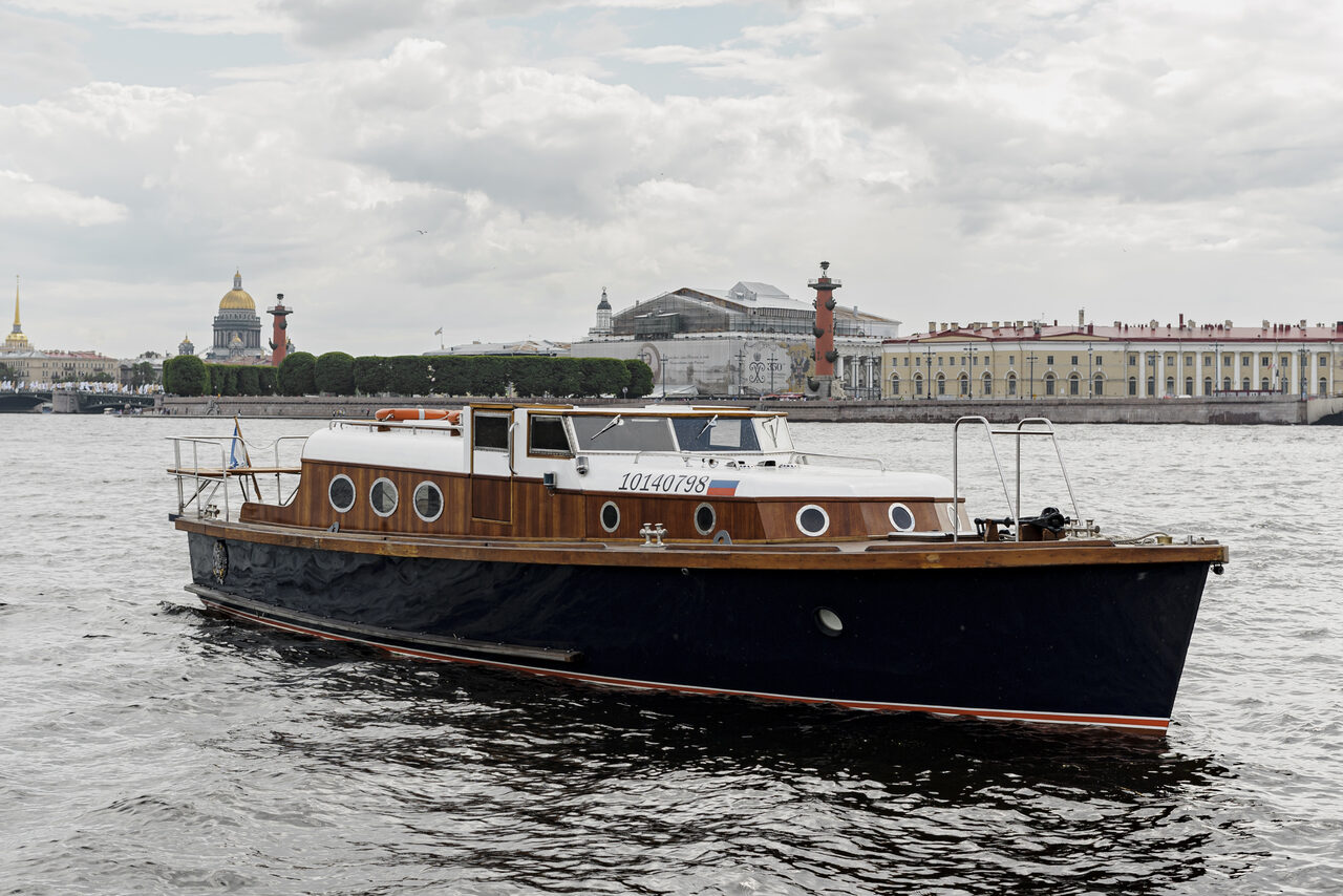 Арендовать Адмиральский катер. Лодка на воде с деревянной палубой и черным корпусом. На фоне городские здания и колонны. Облачно. У лодки круговые окна и открытая верхняя часть с ограждением. На борту текст с цифрами.
