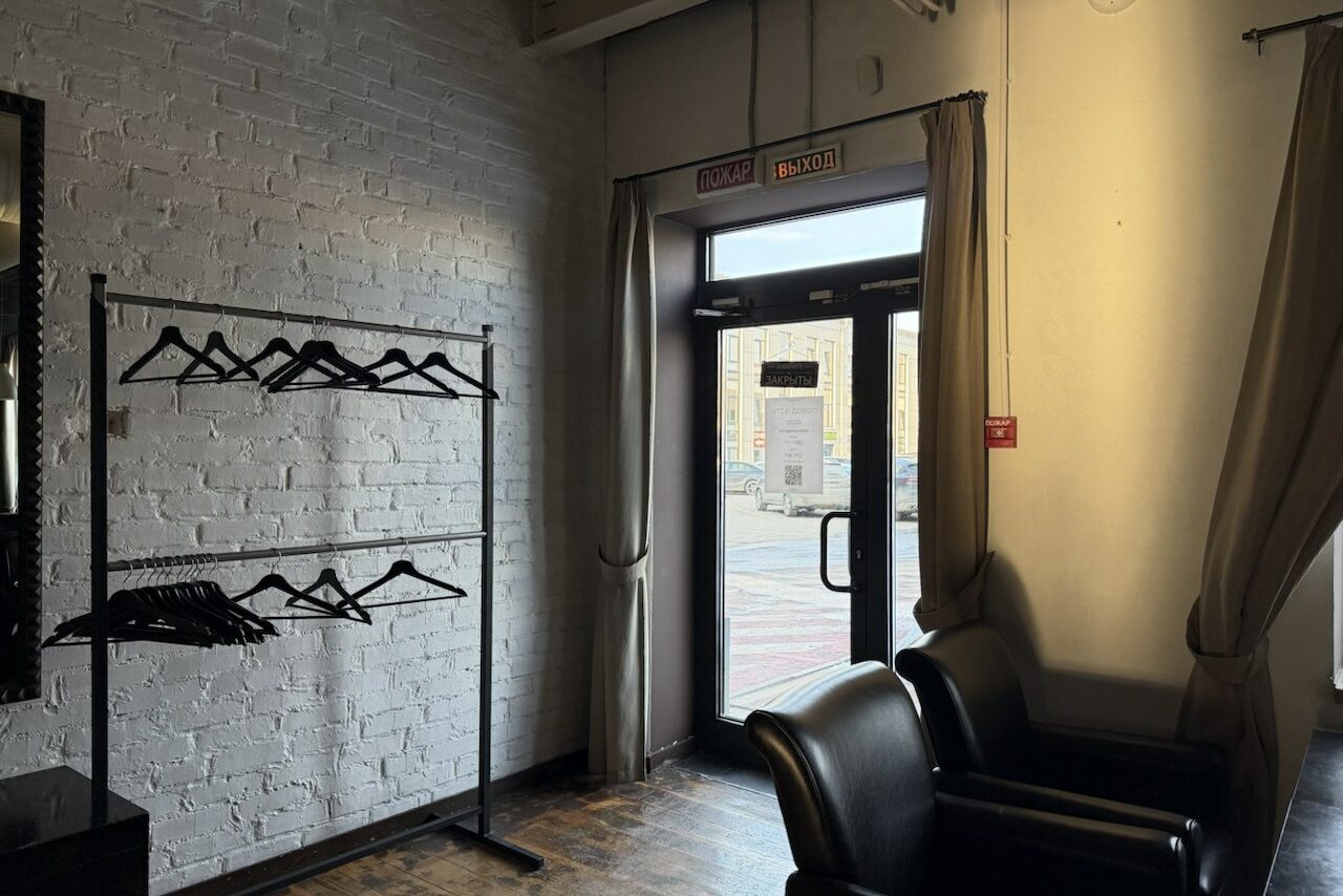 Арендовать Лофт Повод Есть. Стойка с вешалками у белой кирпичной стены. Двойные кожаные кресла справа. Темный деревянный пол. Дверь со стеклянной вставкой, закрытая шторами, надписи "Вход" и "Выход" над дверью. Помещение освещено естественным светом.