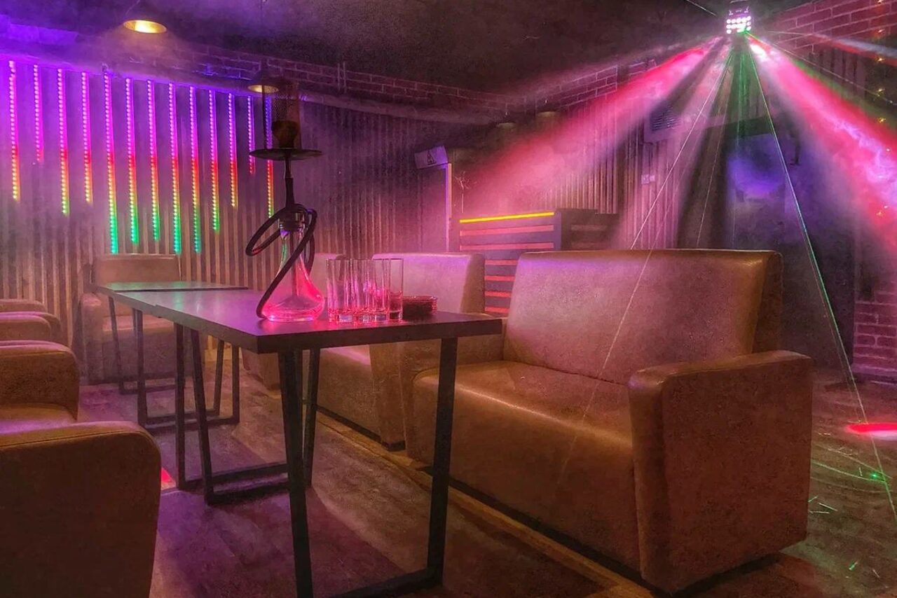 Арендовать Forest(Лес). Кафе с коричневыми диванами, высоким столом, кальяном и бокалами. Вертикальные светодиодные панели на стене, разноцветные световые эффекты. Деревянный пол, кирпичная стена, приглушенное освещение.