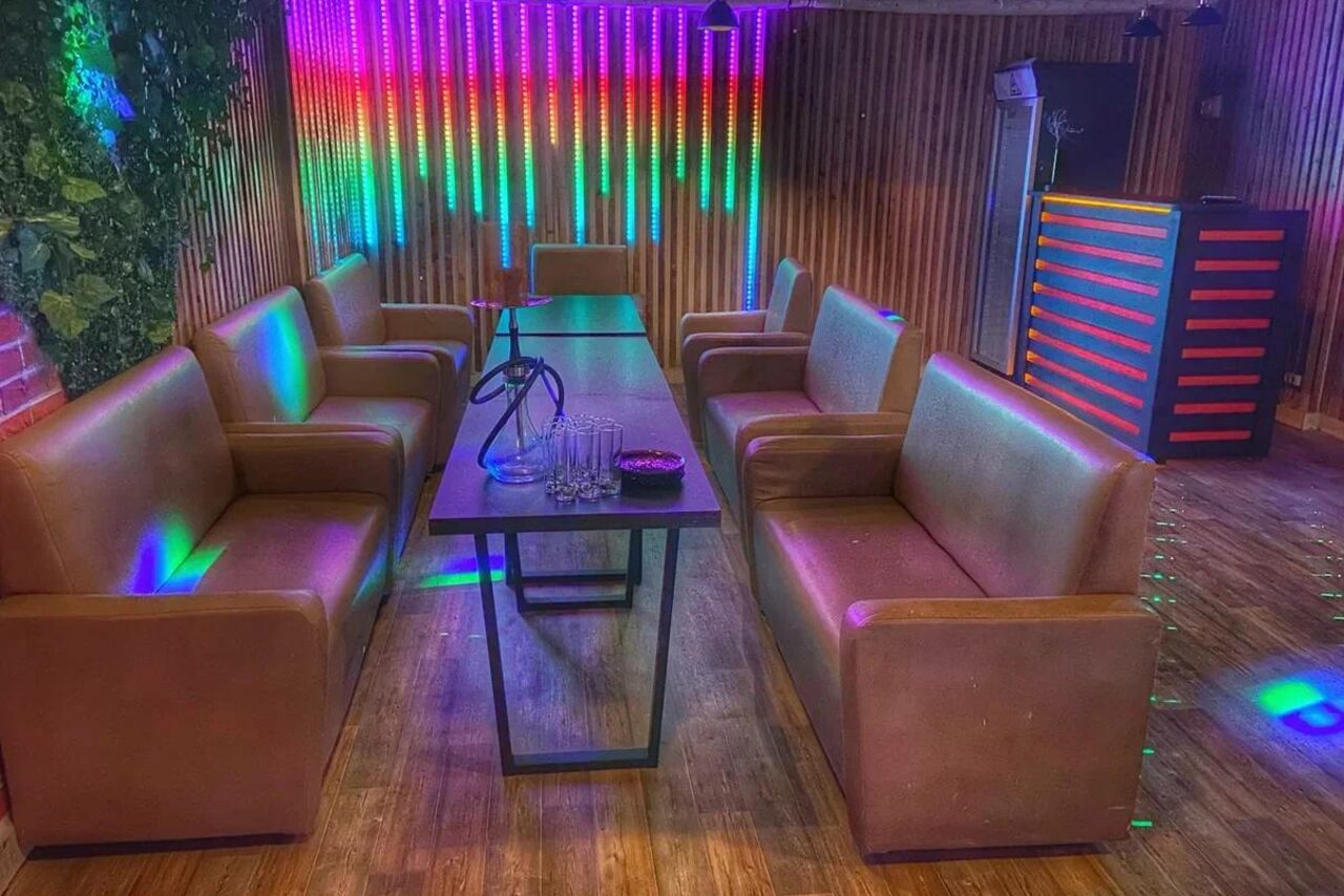 Арендовать Forest(Лес). Современное помещение с диванами из коричневой кожи по обе стороны длинного стола. Вертикальные деревянные панели на стенах. Яркие светодиодные светильники разноцветно освещают пространство. На столе кальян и стеклянные стаканы. Напольное покрытие из дерева.