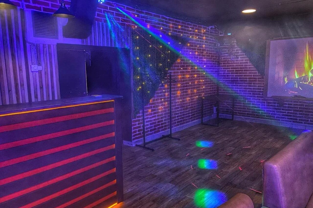 Арендовать Forest(Лес). Темное помещение с кирпичными и деревянными стенами, барной стойкой слева, дискотечными световыми эффектами на полу и стенах, искусственным камином с изображением огня на экране. Потолочные светильники, пустая сцена, паркетный пол.