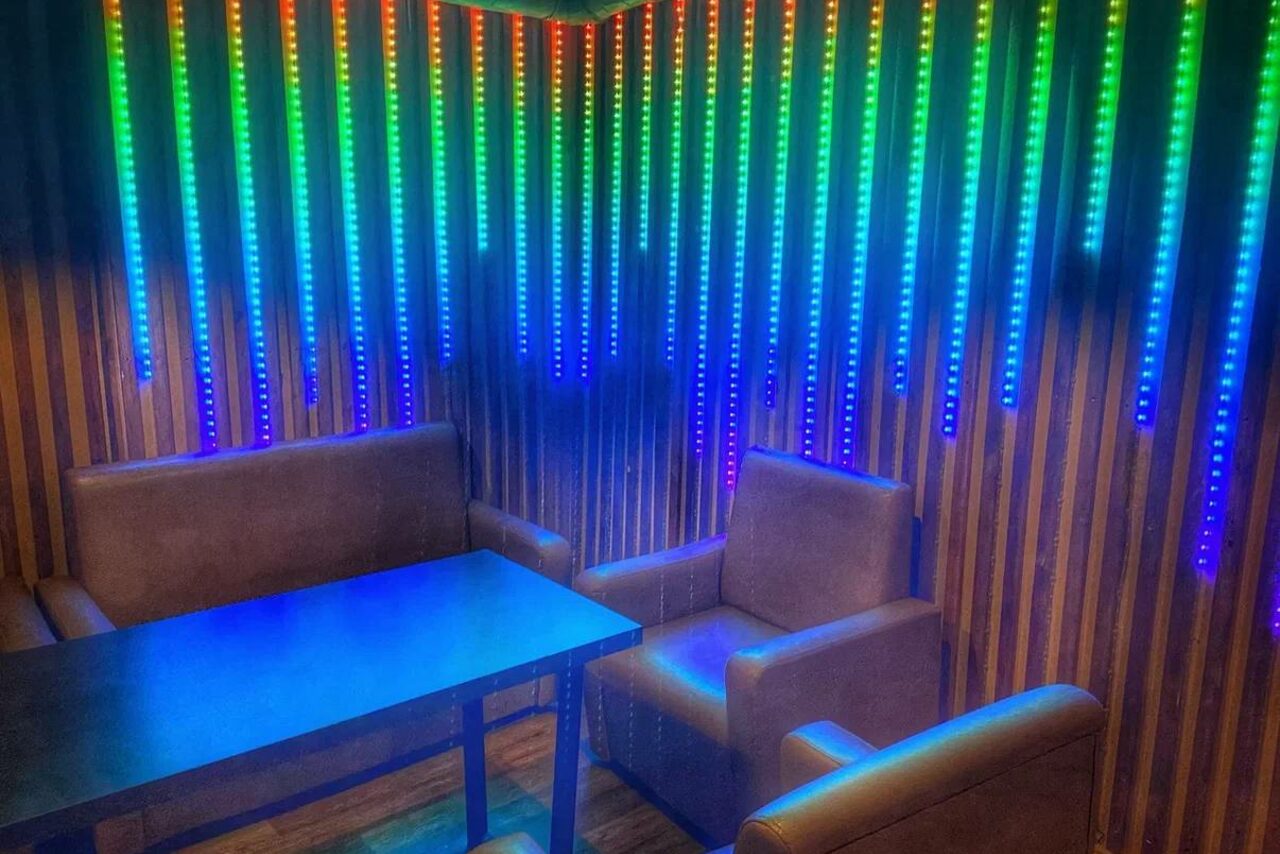 Арендовать Forest(Лес). Современное помещение с мягкими креслами и диваном вокруг прямоугольного стола. Декоративная панель с вертикальными светодиодными лампами, меняющими цвета. Отделка стен деревянными панелями. Пол темный, освещение приглушенное.