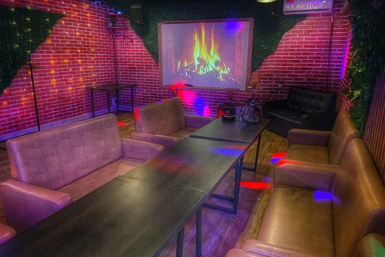 Арендовать Forest(Лес). Караоке-зал с кирпичными стенами, диванами, столом и проектором с изображением камина. На полу деревянное покрытие, освещение разноцветное, стены украшены зеленью.