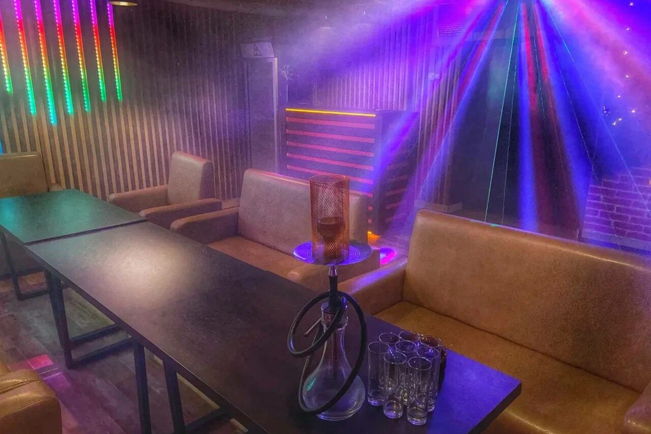 Арендовать Forest(Лес). Тёмное помещение с диванами и столами, на столе кальян и стаканы. Вертикальные деревянные панели на стене, яркие разноцветные светодиодные лампы, фиолетовые световые прожекторы. Пол из тёмного дерева, кирпичная стена.