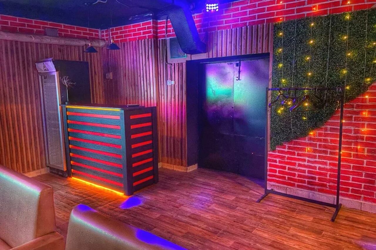 Арендовать Forest(Лес). Помещение с деревянным полом и стенами, оформленными декоративным кирпичом и панелями. Небольшая стойка с подсветкой. Декоративная зелень с огоньками на стене. Есть металлический стеллаж. Мягкие стулья, верхнее освещение, цветное освещение.