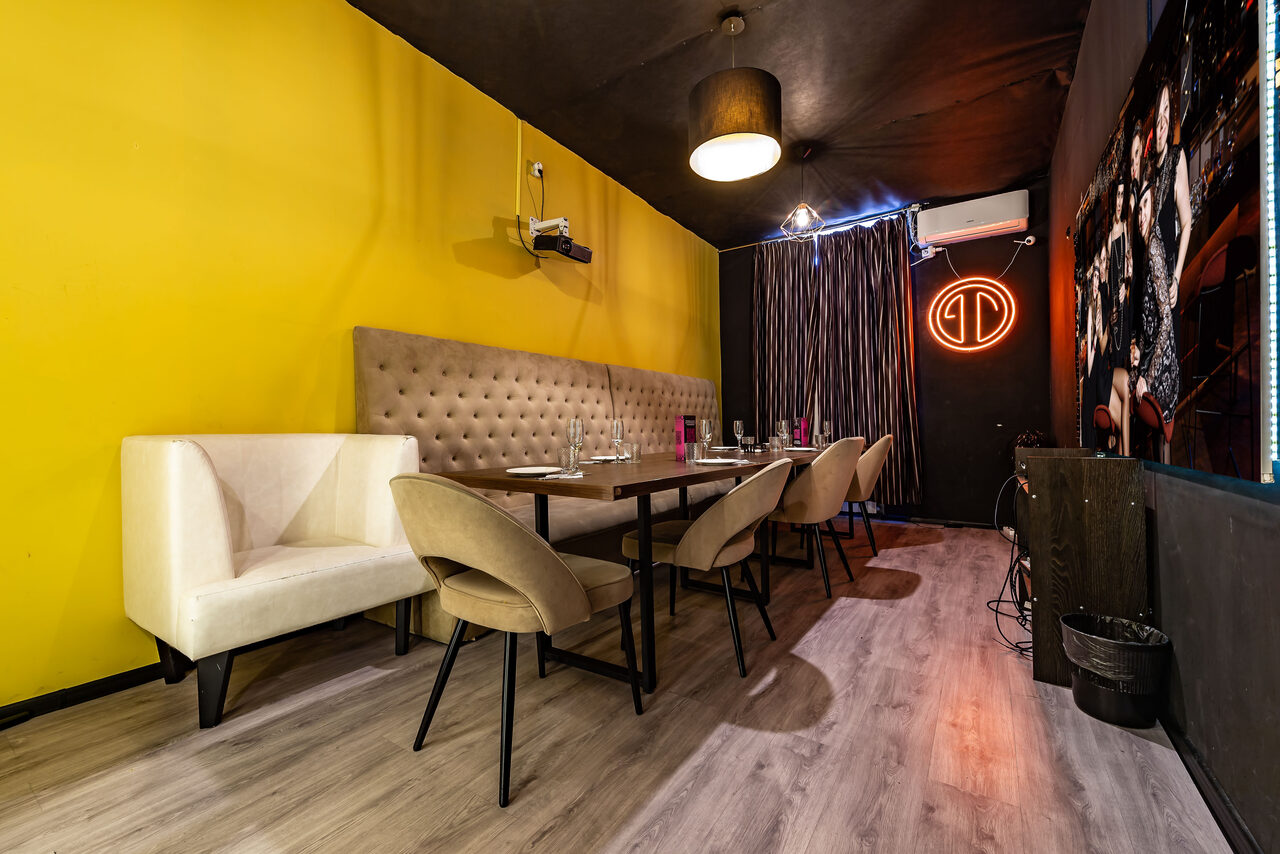 Арендовать Rubin Loft (Рубин Лофт). Зал Гэтсби. Частная комната для мероприятий с белым диваном и бежевыми стульями вокруг деревянного стола. Желтая стена, черный потолок, деревянный пол, подвесной светильник и неоновая вывеска на темной стене. Шторы и кондиционер в углу.