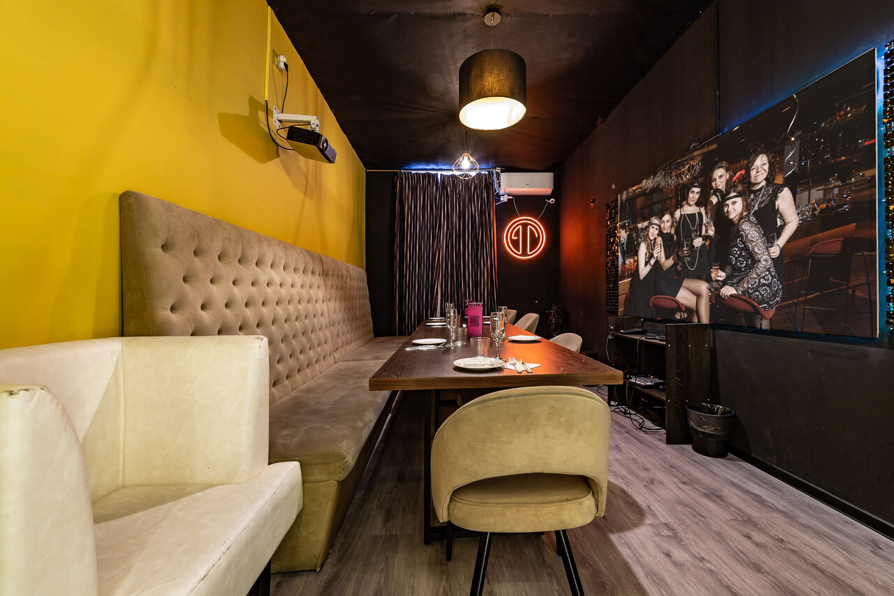 Арендовать Rubin Loft (Рубин Лофт). Зал Гэтсби. Частная комната с длинным столом, обитым диваном и креслами. На стенах постер с изображением людей и неоновый знак. Потолочный светильник, проектор, занавески, кондиционер. Стены желтого и черного цвета, пол светлый.