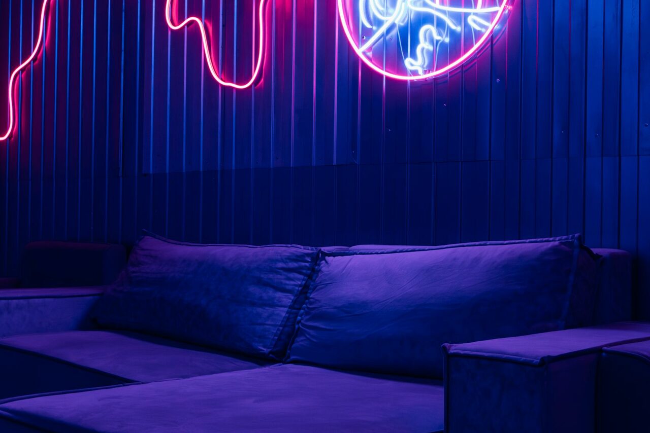 Арендовать Rubin Loft (Рубин Лофт). Зал Токио. Современное помещение с мягким диваном, темно-синими подушками и настенным неоновым светом розовых и синих оттенков. Стены отделаны вертикальными панелями, создающими атмосферу вечернего освещения.