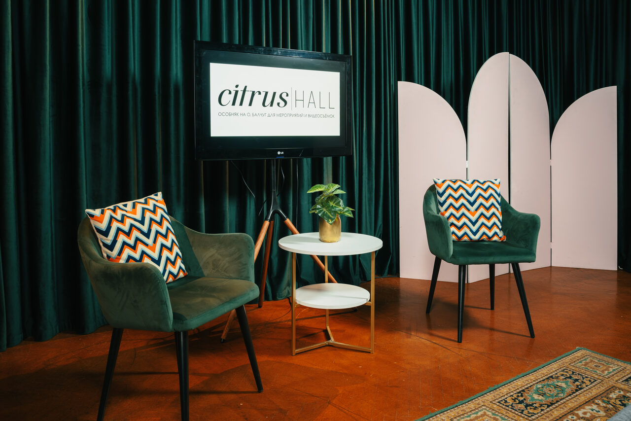 Арендовать Citrus Hall (Цитрус Холл). Зал Мандарин. Зона отдыха с двумя зелёными креслами с узорными подушками, белый круглый стол, на котором стоит растение. На заднем плане тёмно-зеленые занавески, экран с текстом и розовая ширма. Пол коричневый, присутствует ковёр.