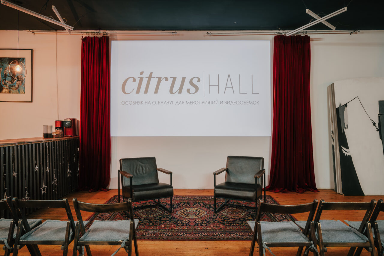 Арендовать Citrus Hall (Цитрус Холл). Зал Грейпфрут. Пространство с деревянным полом, двумя черными креслами и несколькими складными стульями. Красные шторы по бокам экрана с надписью. Черный барный уголок и ковер с узором. Настенное освещение и серые потолочные балки.
