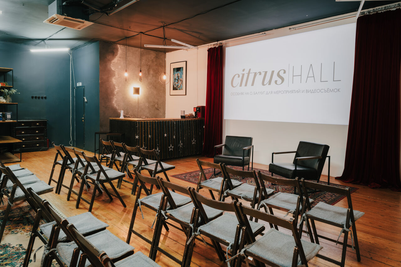 Арендовать Citrus Hall (Цитрус Холл). Зал Грейпфрут. Конференц-зал с деревянным полом и складными стульями, два кожаных кресла у экрана с надписью "citrus HALL". Барная стойка на фоне, серые бетонные стены, синяя акцентная стена, декоративные светильники, бордовые шторы.