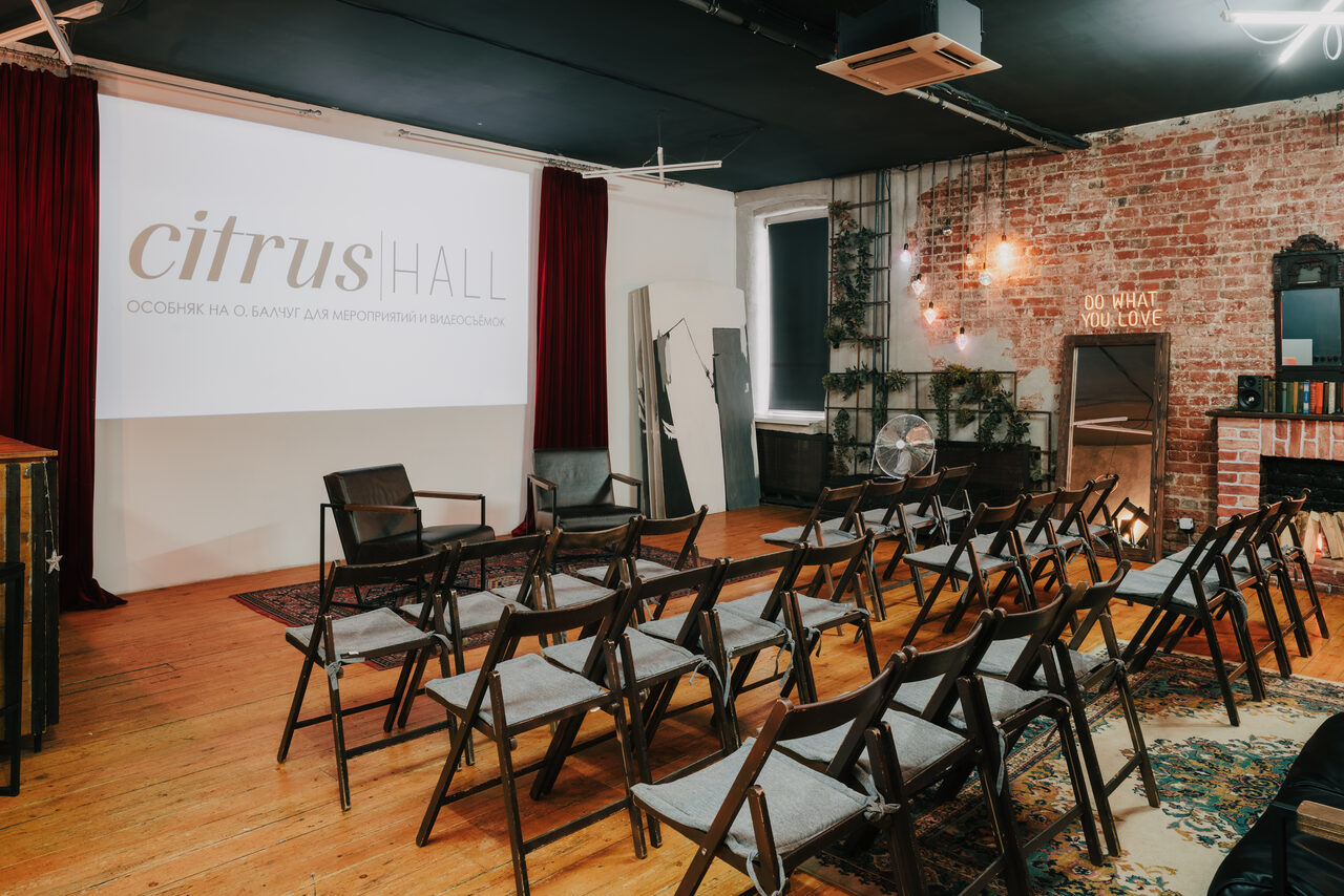 Арендовать Citrus Hall (Цитрус Холл). Зал Грейпфрут. Пространство для мероприятий с экраном на стене, проектор не виден. Ряды складных стульев, деревянный пол, красный занавес слева, кирпичная стена справа с гирляндами. Есть зеркало, вентилятор и книжный шкаф. На стене надпись "DO WHAT YOU LOVE".