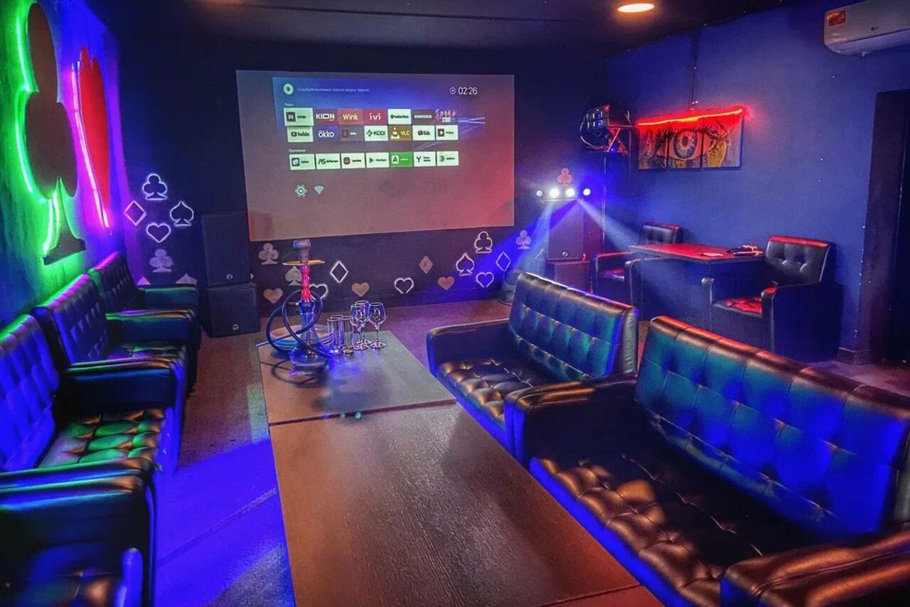 Арендовать Rave(Рейв). Караоке-зал в стиле лаундж с кожаными диванами, низким столом и кальяном. Проектор на стене с интерфейсом меню, разноцветная неоновая подсветка, настенные рисунки с картами, черные стены и полы, фоновое освещение, кондиционер вверху.