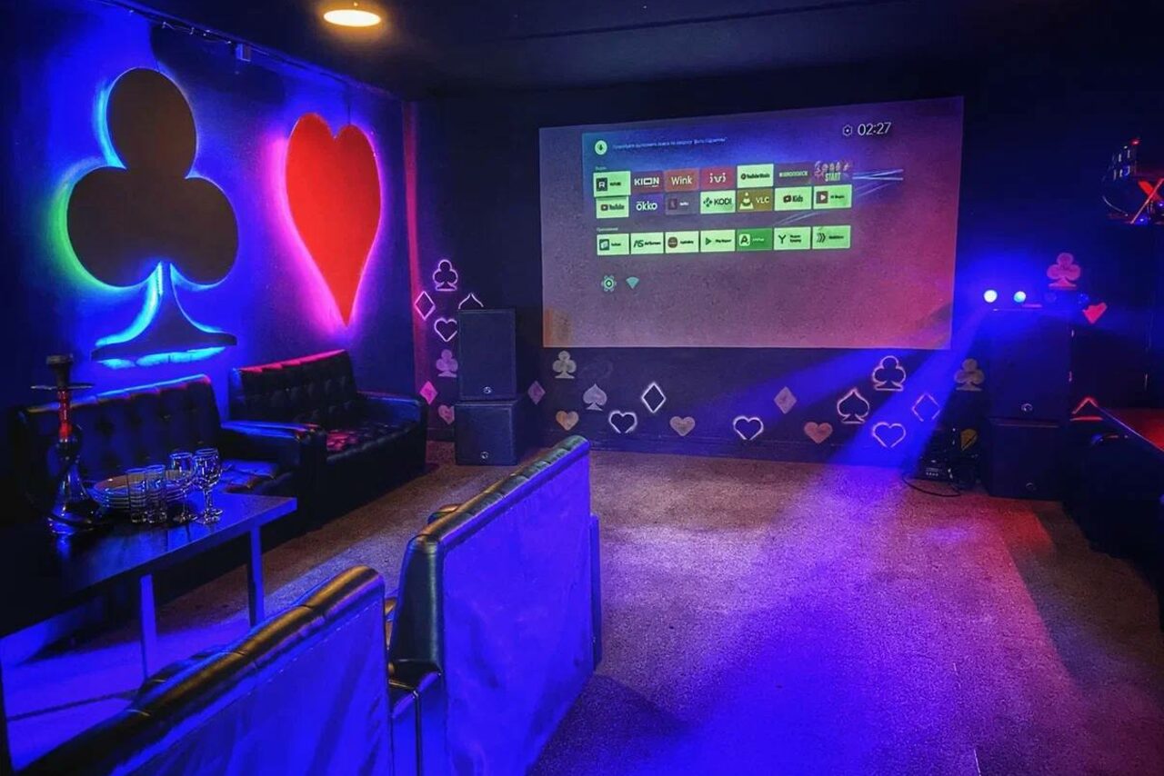 Арендовать Rave(Рейв). Караоке-зал с мягкими кожаными диванами, столом, кальяном. На стене проекционный экран, вокруг которого светящиеся символы мастей карт. Яркая неоновая подсветка в цветах синего и красного. Тёмные стены, ковровое покрытие.