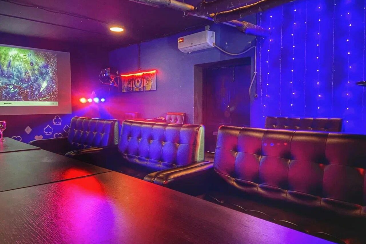 Арендовать Rave(Рейв). Караоке-комната с черными кожаными диванами, деревянными столами и светодиодной подсветкой. На одной стене проектор с изображением, на другой - неоновая подсветка и рисунки. Потолочные споты и гирлянда с синими огнями. Кондиционер на стене.