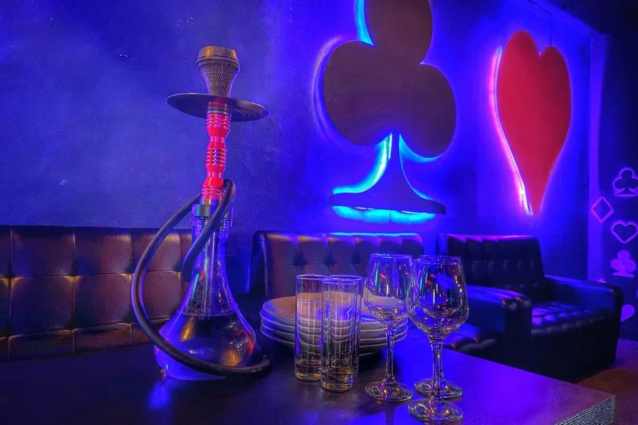 Арендовать Rave(Рейв). Кальянная зона с мягкими черными диванами у стены. На столе кальян, стаканы, бокалы и тарелки. Сине-красная неоновая подсветка в форме мастей игральных карт на стене. Атмосферное освещение, тёмные стены и мебель.