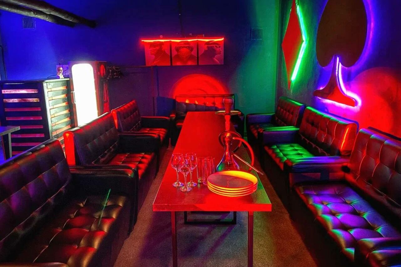 Арендовать Rave(Рейв). Караоке-зал с красной мебелью: длинный стол, кожаные диваны. На столе стеклянная посуда и кальян. Неоновое освещение в виде карточных мастей. Стены темные. Наличие холодильника. Настенное изображение из трех сегментов.