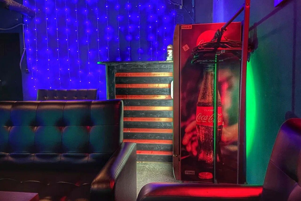 Арендовать Rave(Рейв). Кожаные диваны, стойка с красными полосами, холодильник с изображением бутылки, фиолетовая светодиодная гирлянда на стене, разноцветное освещение; пространство с элементами декора в тёмных и ярких цветах.