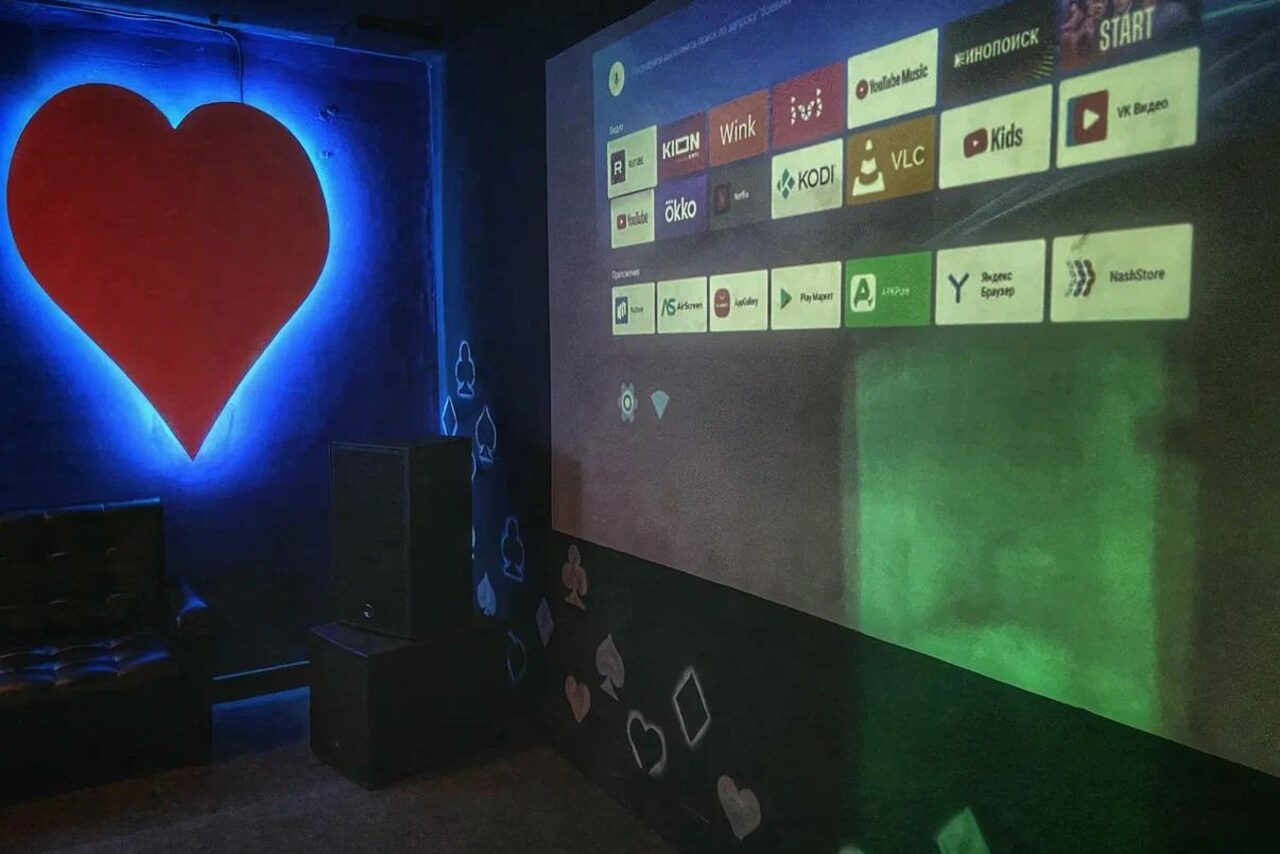 Арендовать Rave(Рейв). Комната с темными стенами, декорированная изображениями карт и светящимися элементами; на одной из стен проецируется экран с меню приложений. Слева кожаное кресло и большая колонка, на стене светится красное сердце. Светится синий контур.