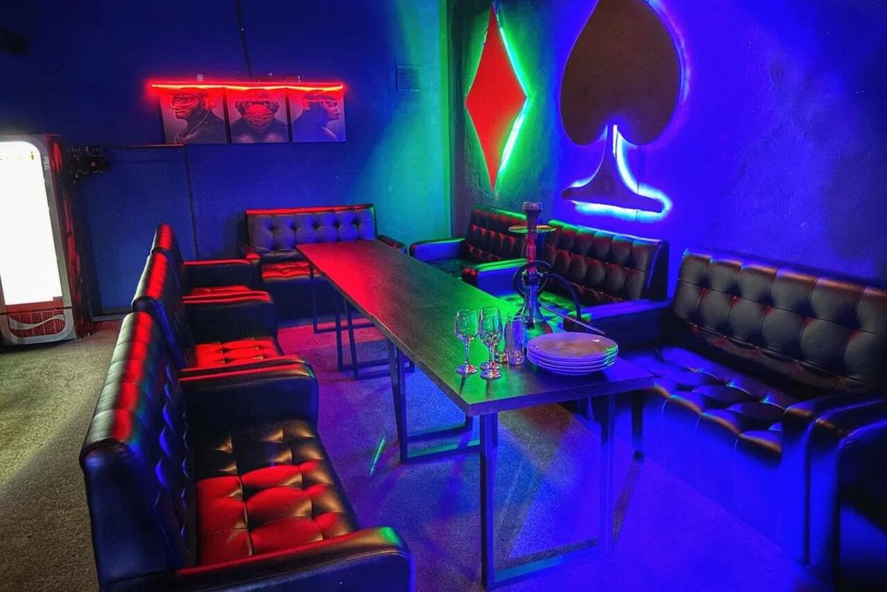 Арендовать Rave(Рейв). Бар сине-зелёной подсветкой, чёрные кожаные диваны и стулья, длинный стол с бокалами и тарелками, настенные неоновые фигуры карты и красная гирлянда. На стене плакат с изображениями. Справа холодильник.