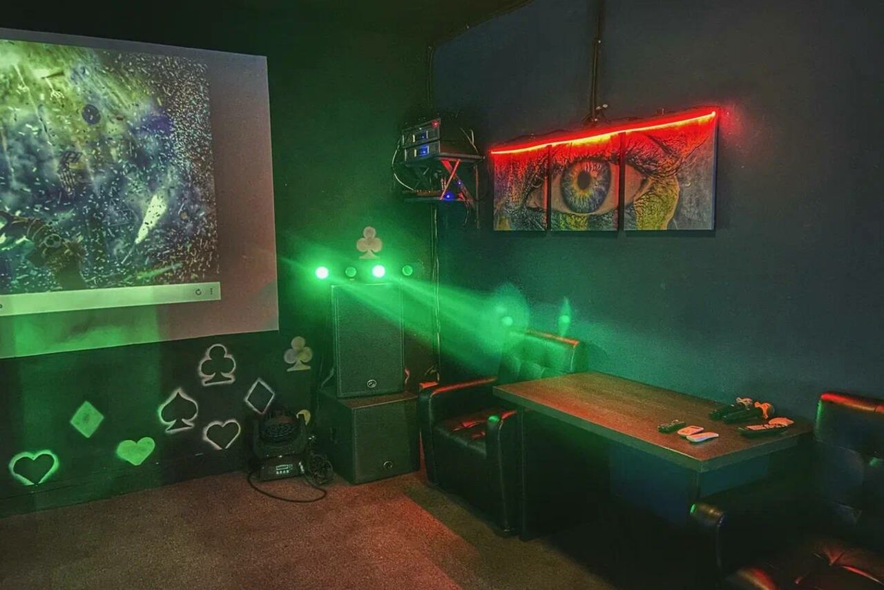 Арендовать Rave(Рейв). Комната с затемнённым освещением, мебель: кожаные кресла, стол. На стене проектор с изображением, граффити с карточными мастями и настенная картина с изображением глаза. Звуковая аппаратура, световая инсталляция с зелёными лучами. Тёмные стены и ковёр.