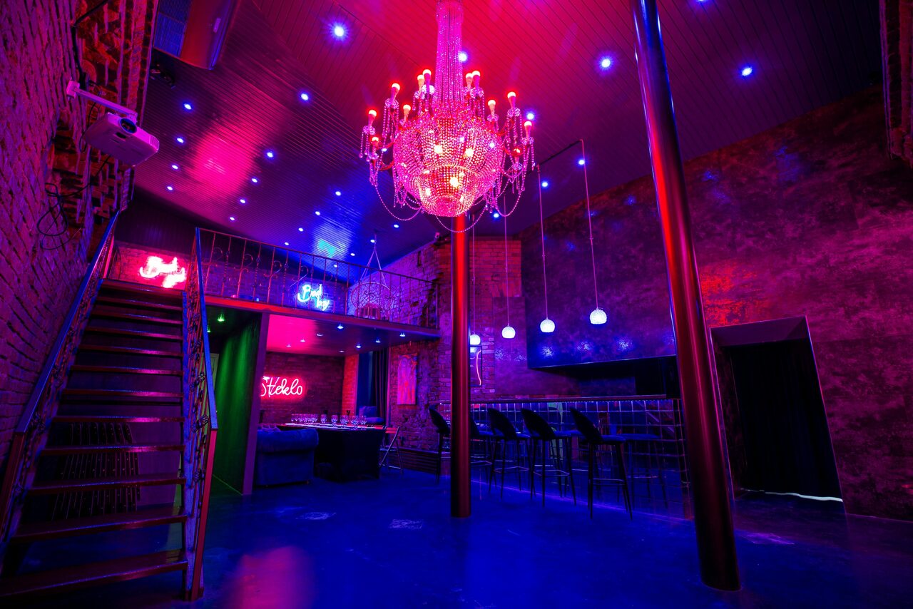 Арендовать Двухэтажный лофт с отдельным входом. Интерьер помещения с лестницей и барной стойкой. Красные и синие неоновые огни, хрустальная люстра, кирпичные стены. Подвесные светильники над барной стойкой с высокими стульями. Металлические перила и бетонные элементы. Имеются яркие неоновые надписи.