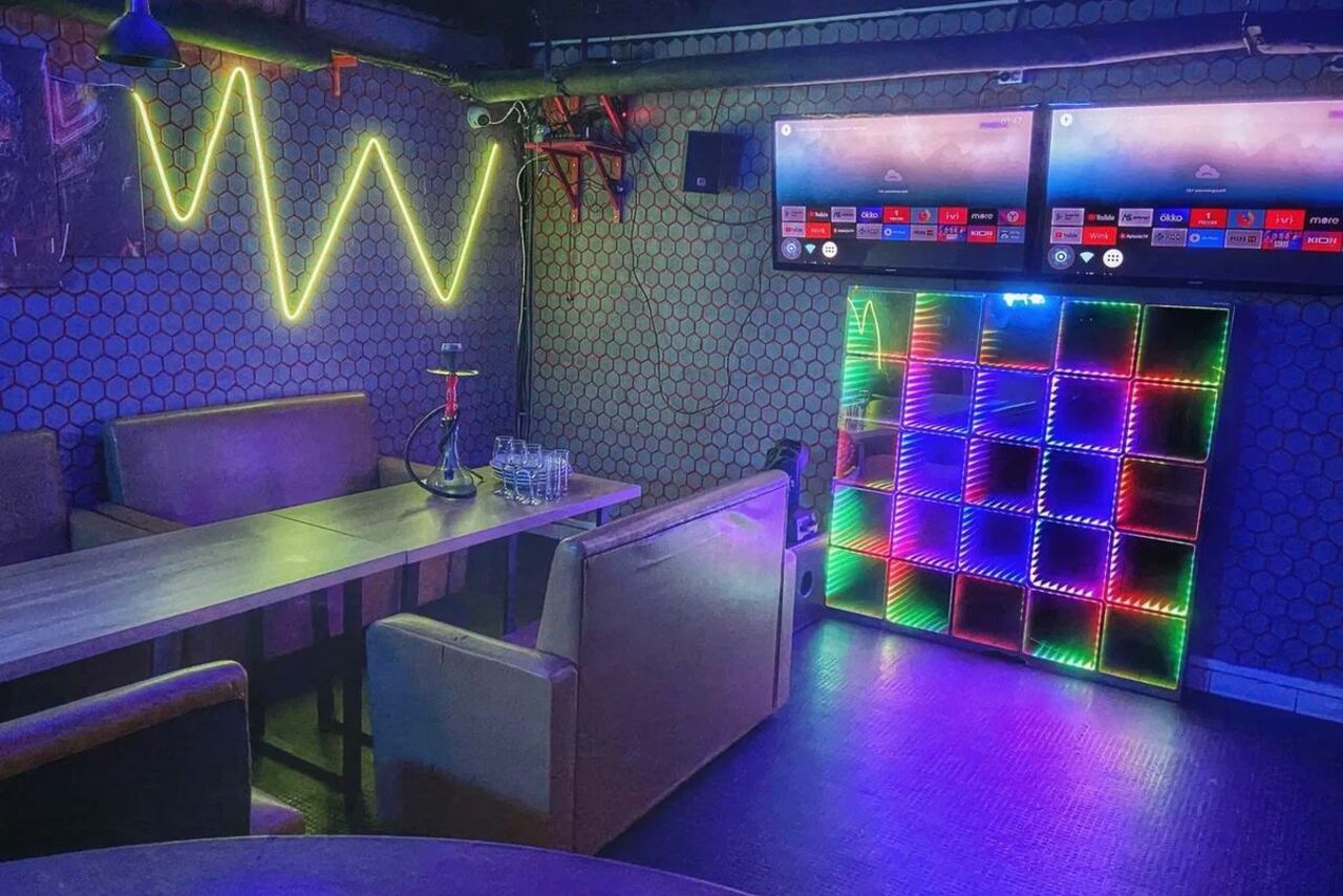 Арендовать Bee-lounge(Пчела-лаундж). Частное помещение с мягкими диванами и длинным столом, два настенных телевизора, неоновая подсветка в виде линий и цветная многоярусная панель, кальян на столе, стены с геометрическим узором, тёмный пол.