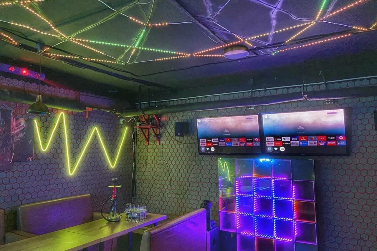 Арендовать Bee-lounge(Пчела-лаундж). Караоке-зал с двумя телевизорами на стене, рядом стол с диваном. На стенах и потолке светодиодные ленты разных цветов, неоновая надпись, шестигранная плитка. На столе стеклянные стаканы и кальян. Освещение разноцветное.