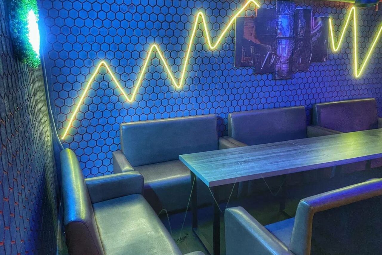 Арендовать Bee-lounge(Пчела-лаундж). Частная зона отдыха с кожаными диванами и прямоугольным деревянным столом. Стены декорированы синими шестиугольниками и неоновыми зигзагообразными лампами. Настенное изображение в темных тонах, приглушенное освещение.