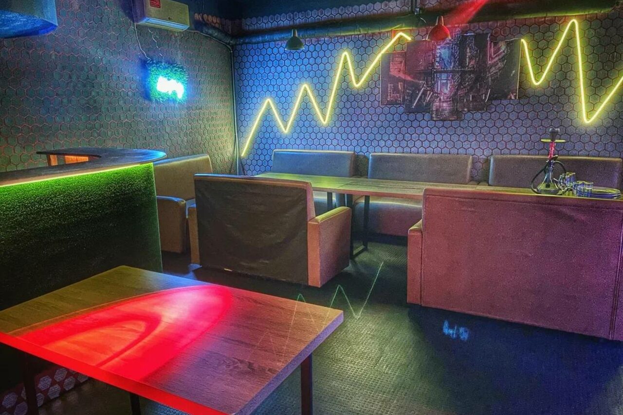 Арендовать Bee-lounge(Пчела-лаундж). Лофт-пространство с диванами и столами, неоновая подсветка на стене с рисунком линий, декорирована граффити, на столе кальян. Тёмный пол, стены с узором из шестиугольников, разноцветное освещение, зелёный неоновый знак.