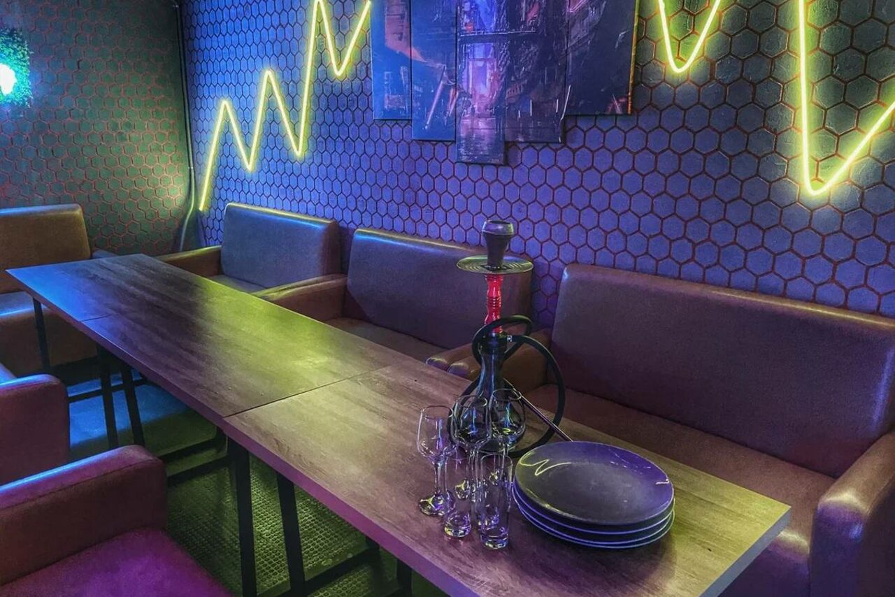 Арендовать Bee-lounge(Пчела-лаундж). Комната с длинным обеденным столом и коричневыми диванами вдоль стены. На столе кальян, тарелки и бокалы. Стена декорирована геометрическими обоями и неоновым освещением в виде диаграммы. На стене несколько картин.