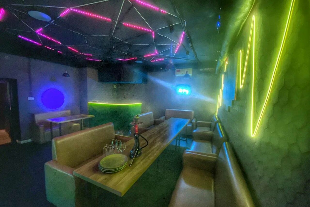 Арендовать Bee-lounge(Пчела-лаундж). Лофт-пространство с неоновым освещением. В центре длинный стол с мягкими диванами по бокам. На столе стеклянная посуда и кальян. Потолок с геометрическим узором из неоновых полос. Стены с неоновыми акцентами.