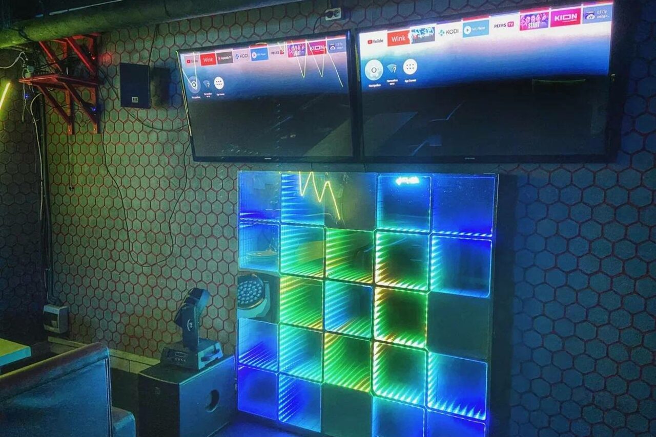 Арендовать Bee-lounge(Пчела-лаундж). Клубный зал с двумя настенными экранами, ярким разноцветным светодиодным панно и оборудованием для звука. Стена с серым шестигранным узором. Помещение с приглушенным освещением, проводами на стене и мебелью для сидения.