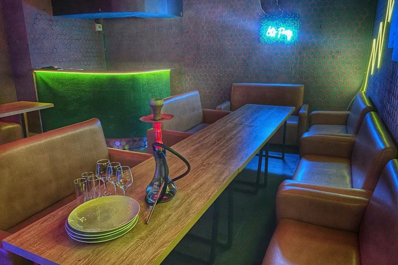 Арендовать Bee-lounge(Пчела-лаундж). Комната с длинным деревянным столом, окружённым кожаными креслами. На столе стеклянные бокалы, тарелки и кальян. На стене неоновая надпись. Стены обиты тёмными материалами, подсвеченный бар в углу. Освещение разноцветное.