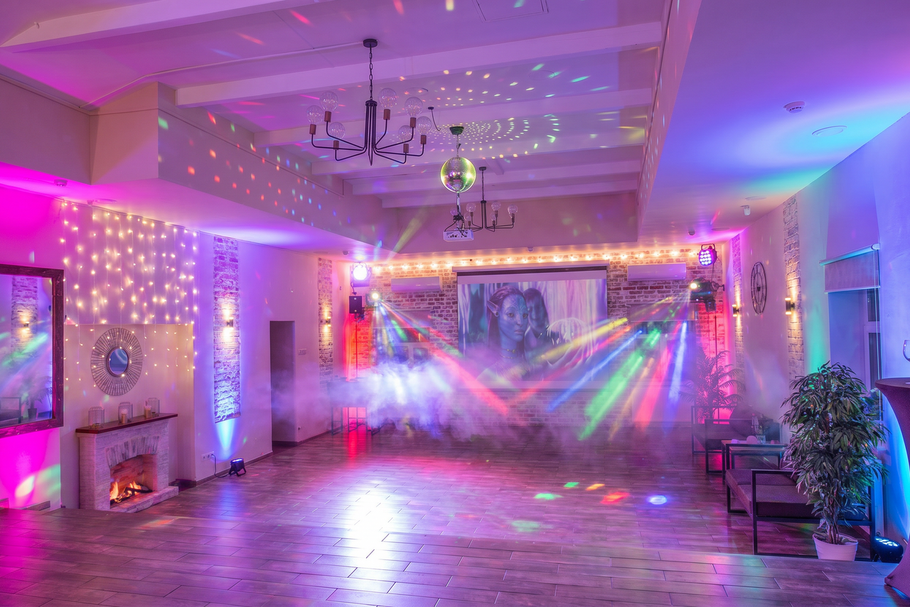 Арендовать Loft Lodge (Лофт Лодж) 7. Просторный уютный лофт. Просторное помещение с деревянным полом и белым потолком. Декоративный камин с подсветкой, на стенах кирпичная отделка. Светодиодные гирлянды и разноцветные прожекторы создают атмосферу. Есть экран с проектором, стулья, растения. На стенах часы и зеркала.