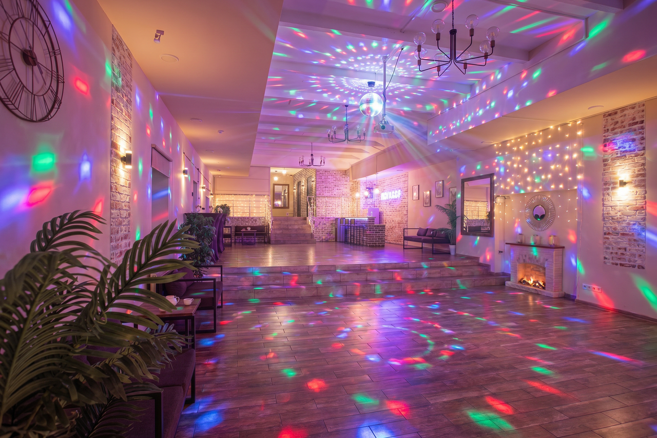 Арендовать Loft Lodge (Лофт Лодж) 7. Просторный уютный лофт. Большое пространство с деревянным полом и кирпичными стенами. На потолке светящиеся гирлянды и диско-шар, проецирующий разноцветные огни. Есть декоративный камин, столы, стулья, зелёные растения и неоновая вывеска. Висит настенные часы.
