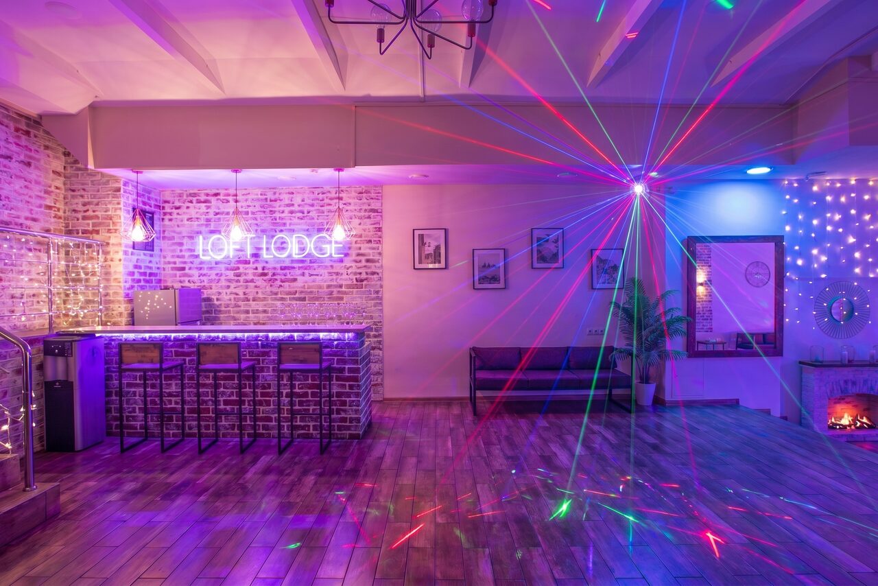Арендовать Loft Lodge (Лофт Лодж) 7. Просторный уютный лофт. Лофт-пространство с барной стойкой из белого кирпича и высокими стульями, кирпичные стены. На стене неоновая надпись «LOFT LODGE». Пол деревянный, разноцветные лазеры, фальш-камин, декоративные лампы, фоторамки.