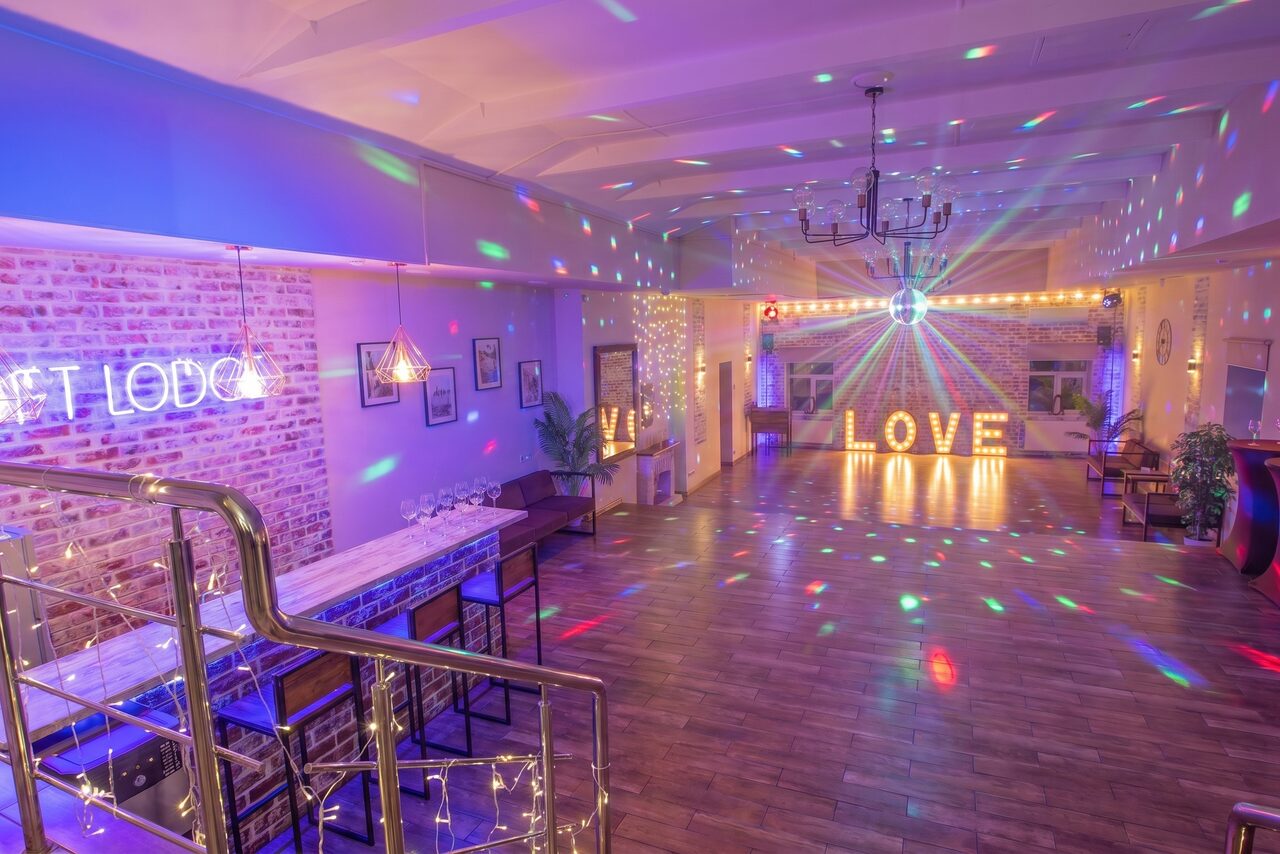 Арендовать Loft Lodge (Лофт Лодж) 7. Просторный уютный лофт. Просторное современное помещение с барной стойкой, стульями и деревянным полом. Мягкое разноцветное освещение, неоновая вывеска на кирпичной стене и большие светящиеся буквы "LOVE". Лестница с металлическими перилами, висячие светильники и декорированные растения.