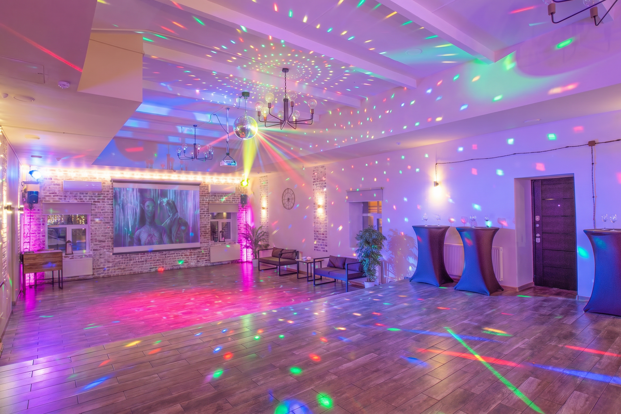 Арендовать Loft Lodge (Лофт Лодж) 7. Просторный уютный лофт. Просторная комната с паркетным полом, диско-шаром и разноцветными световыми эффектами. Стены украшены гирляндами и растениями. У стены расположен проектор, несколько диванов и высоких столов без скатертей. Потолок украшен светильниками и балками.
