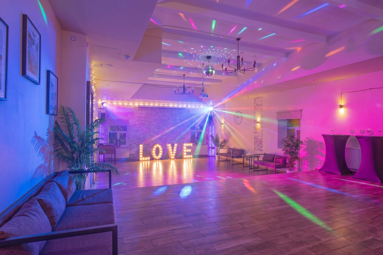 Арендовать Loft Lodge (Лофт Лодж) 7. Просторный уютный лофт. Большое помещение с деревянным полом и кирпичными стенами. Центральное освещение — зеркальный шар и прожекторы. Надпись "LOVE" из светящихся букв. Размещены диваны, растительность, высокий столик. Мягкое разноцветное освещение.