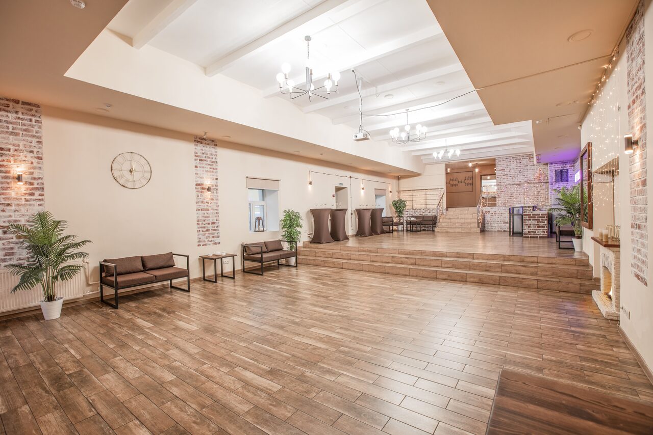 Арендовать Loft Lodge (Лофт Лодж) 7. Просторный уютный лофт. Просторное светлое помещение с деревянным полом и белыми потолочными балками. Стены с отделкой под кирпич и светло-бежевой штукатуркой. Есть коричневые диваны, столики и зеленые растения. Потолочные люстры и настенные светильники.