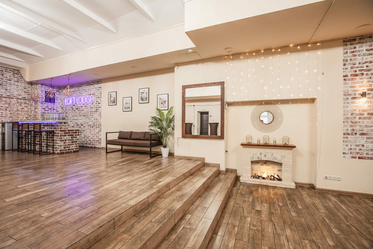 Арендовать Loft Lodge (Лофт Лодж) 7. Просторный уютный лофт. Просторное помещение с деревянным полом, кирпичными стенами и неоновой вывеской. Зона отдыха с диваном и растением. Искусственный камин с декорациями. Светлый интерьер с настенной фотографией и зеркалом. Подвесное освещение.