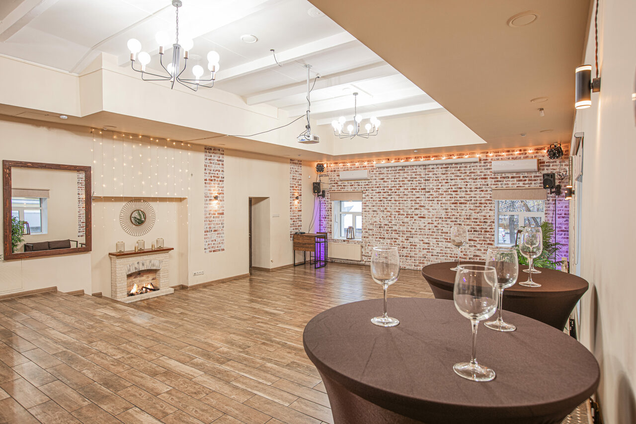 Арендовать Loft Lodge (Лофт Лодж) 7. Просторный уютный лофт. Просторное помещение с кирпичными стенами, деревянным полом и белыми потолками. Декоративный камин, зеркала, круглые столы с бокалами. Большие окна, подвесные светильники и гирлянды с лампочками. Светлые тона интерьера.