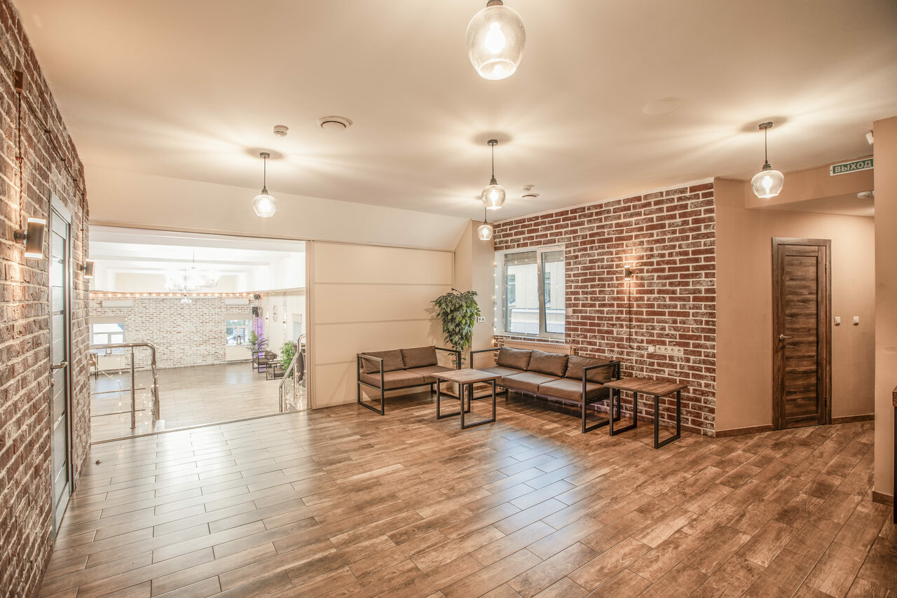 Арендовать Loft Lodge (Лофт Лодж) 7. Просторный уютный лофт. Просторное помещение с кирпичной стеной и деревянным полом, диванами и столиками, растением в углу. Подвесные светильники, табличка с надписью "выход". Окно без занавесок, дверь справа. Современный дизайн, теплое освещение.
