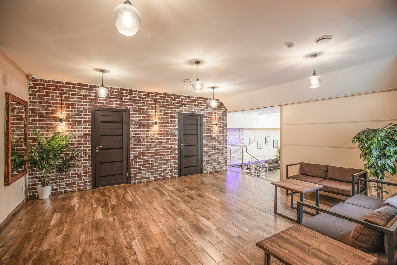 Арендовать Loft Lodge (Лофт Лодж) 7. Просторный уютный лофт. Просторный холл с коричневыми дверями и кирпичной стеной. Пол из деревянных панелей. Мягкая мебель и столики справа. Растения в горшках. Подвесные светильники на потолке. Табличка на стене. Лестница в дальней части. Белые стены слева.
