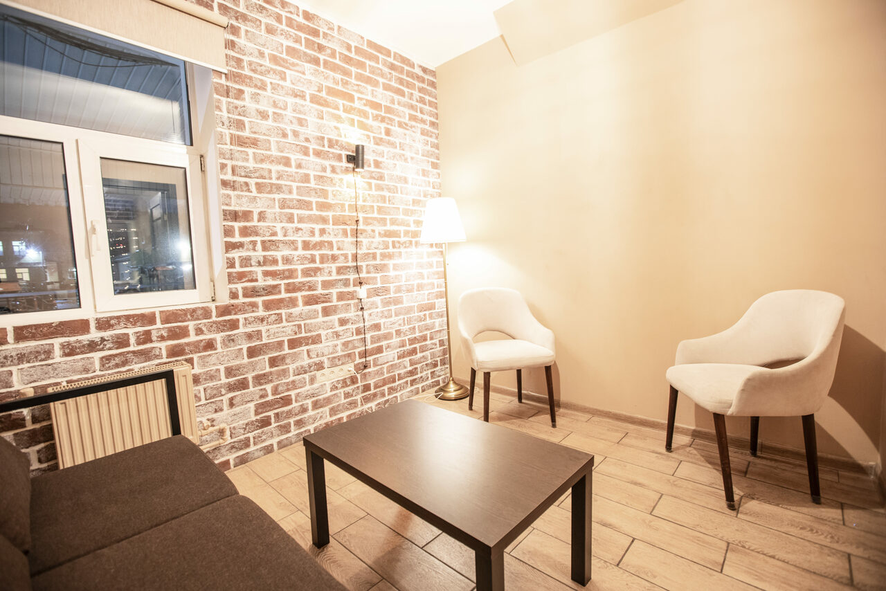 Арендовать Loft Lodge (Лофт Лодж) 7. Просторный уютный лофт. Небольшая комната с кирпичной стеной и бежевыми стенами, двумя белыми креслами, тёмным прямоугольным столом и диваном. Окно с видом на город. Настенное и напольное освещение. Бежевый кафель на полу.