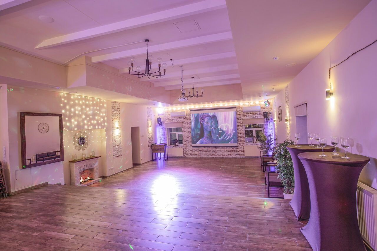 Арендовать Loft Lodge (Лофт Лодж) 7. Просторный уютный лофт. Просторное помещение с паркетным полом, белыми стенами и потолком с балками. На стене установлен проектор. В углу фальшкамин. По бокам расставлены высокие круглые столы с накрытием. Освещение состоит из подвесных светильников и гирлянд.