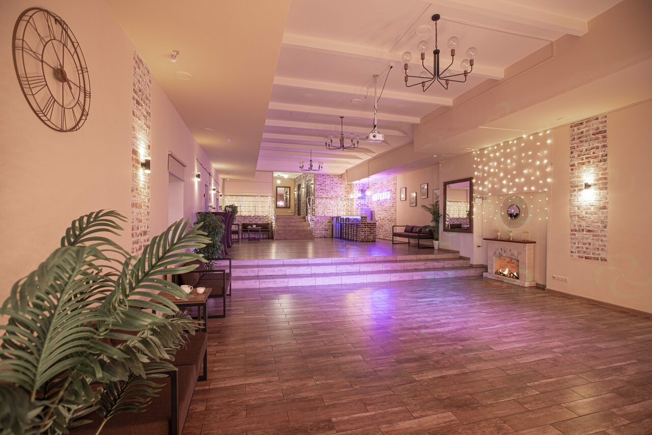 Арендовать Loft Lodge (Лофт Лодж) 7. Просторный уютный лофт. Просторное помещение с деревянным полом, диванами, растениями и камином. На стенах декор в виде кирпичной кладки и струящихся огней. Подвесные светильники и зеркала, часы на стене. Зона с барной стойкой в глубине. Мягкое розовое освещение.